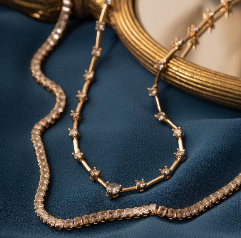 na foto fundo de pano azul e dois colares exuberantes uma riviera dourada com pedras de zirconia e um colar dourado com elo elos de zirconia.